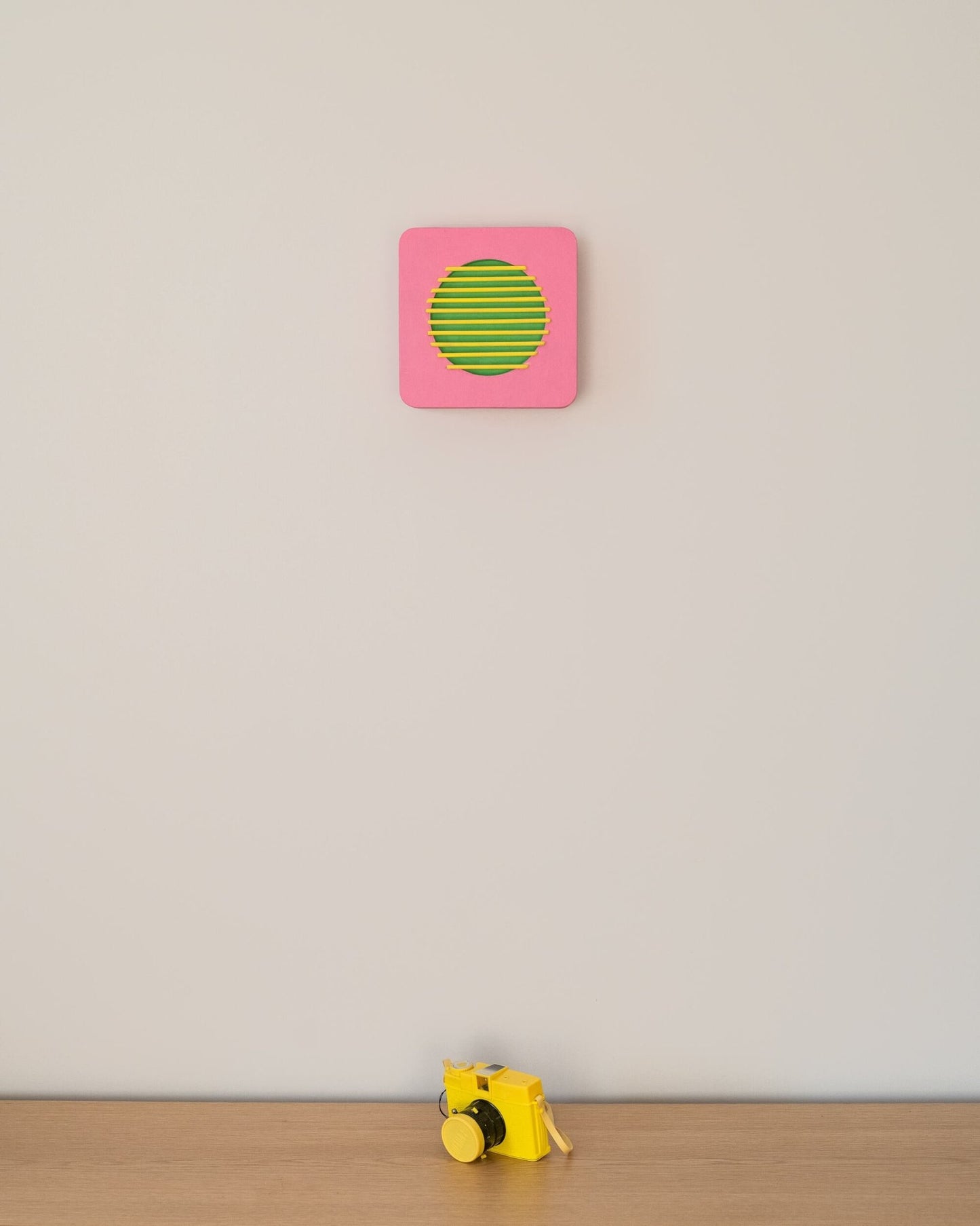 Wall-mounted pink and green geometric sculpture with yellow bars, shown above a small yellow camera for size reference in a minimalist room.