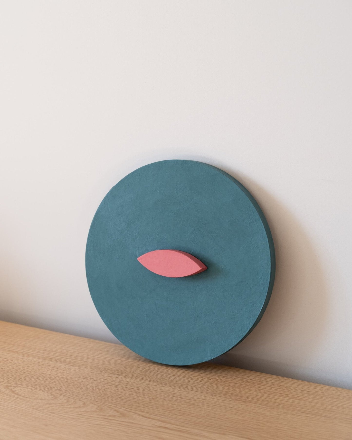Teal circular 3d art object with a pink leaf-like shape on a light wooden surface and white background