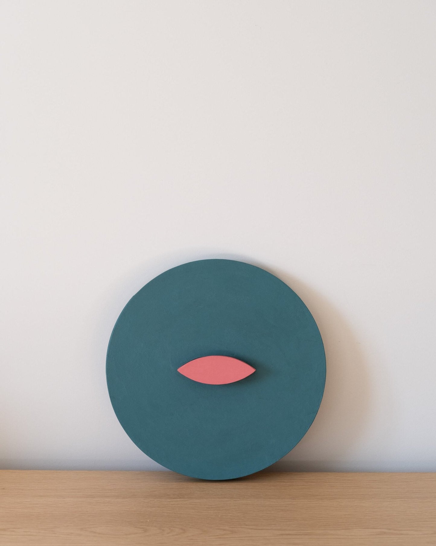 Teal circular wall art object with a pink leaf-like shape on a light wooden surface and white background