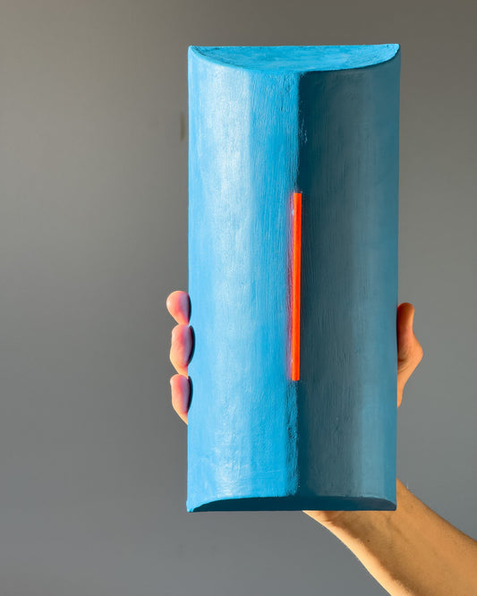 A hand holds a minimalist, geometric blue sculpture with a vertical red-orange accent, designed by Olshbau.