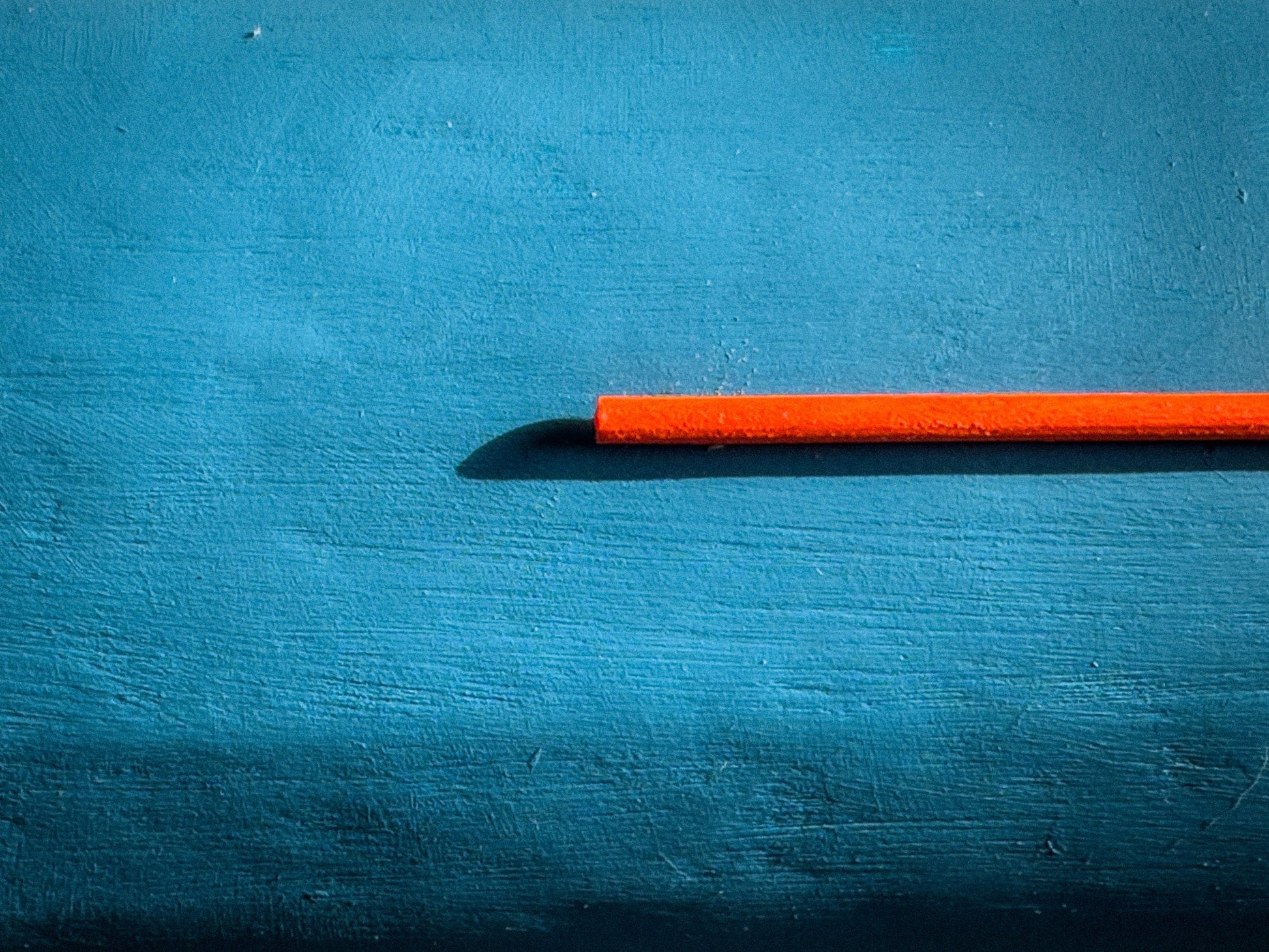 close-up of a blue textured surface featuring a protruding red-orange rod casting a shadow, part of a minimalist geometric sculpture by Olshbau.