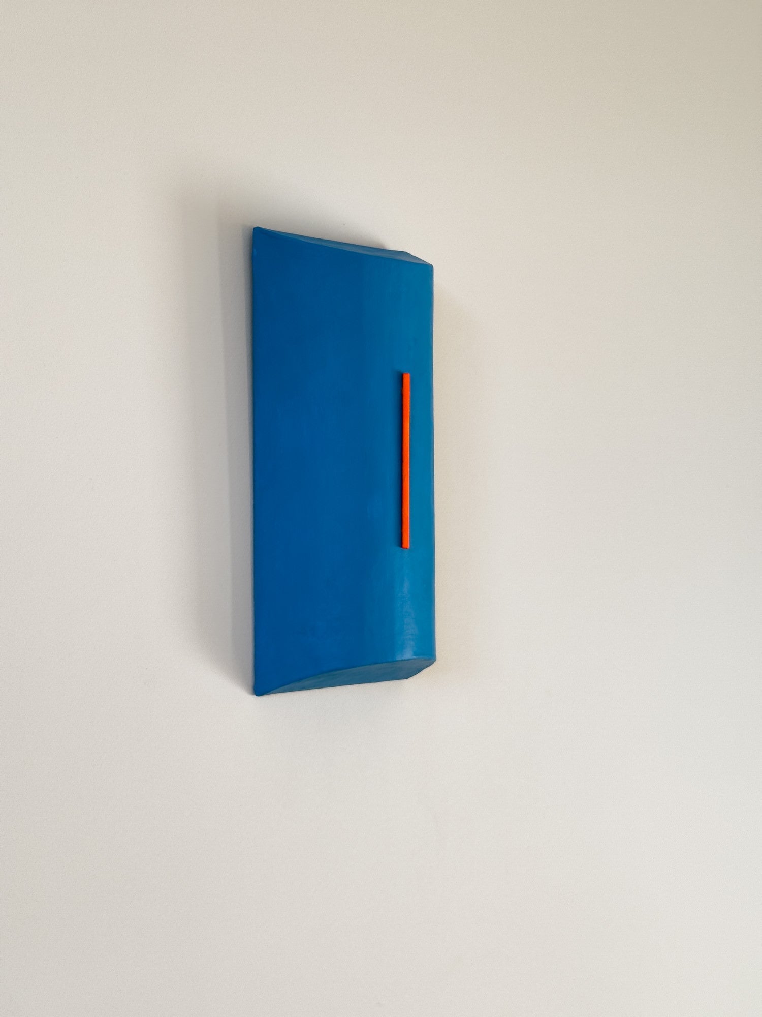 minimalist geometric blue wall sculpture with a vertical red-orange accent by Olshbau, mounted at an angle on a white wall.