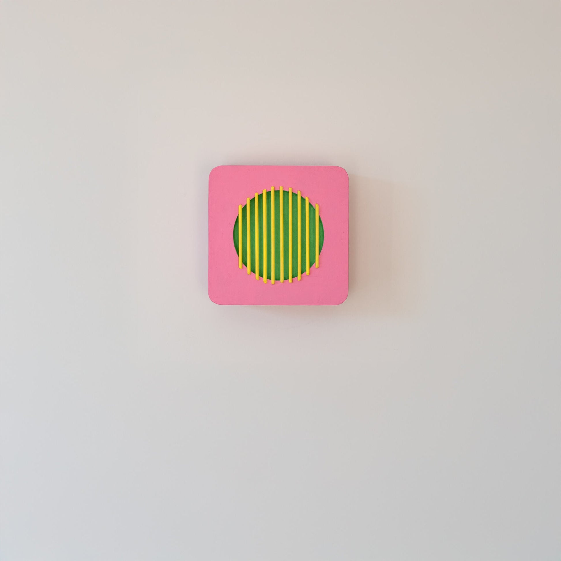 Centered view of a pink square wall sculpture with green circle and yellow bars, displayed on a light wall in minimalist interior.