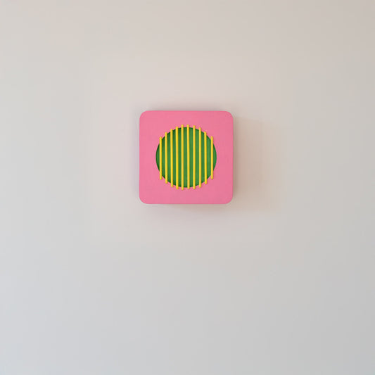 Centered view of a pink square wall sculpture with green circle and yellow bars, displayed on a light wall in minimalist interior.