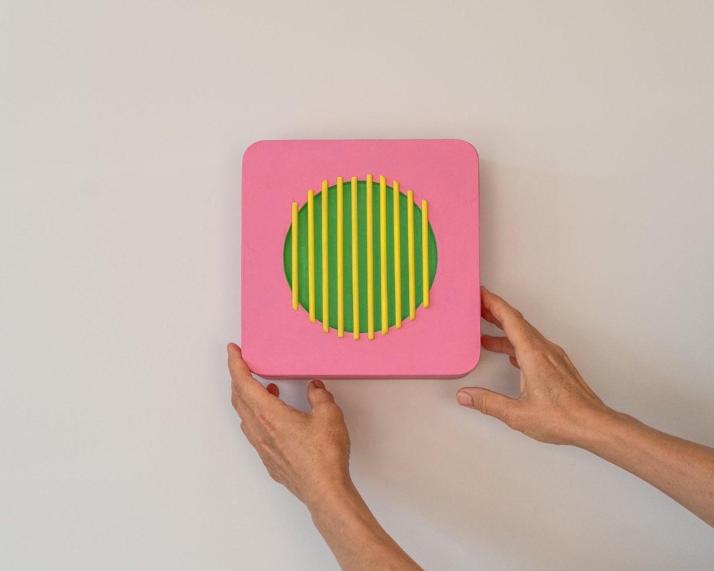 Hands holding a pink and green geometric wall sculpture with yellow bars, demonstrating size and easy wall placement.