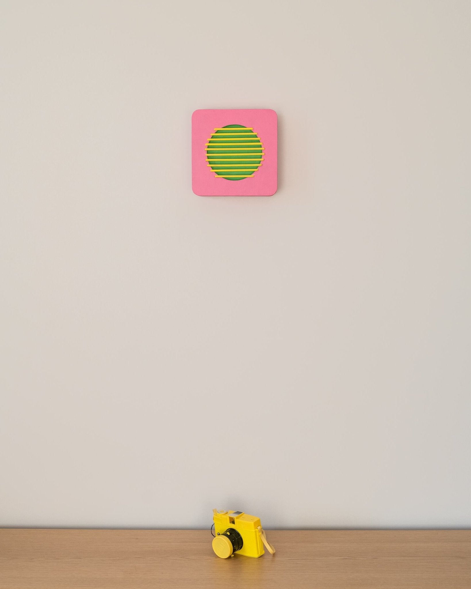 Wall-mounted pink and green geometric sculpture with yellow bars, shown above a small yellow camera for size reference in a minimalist room.