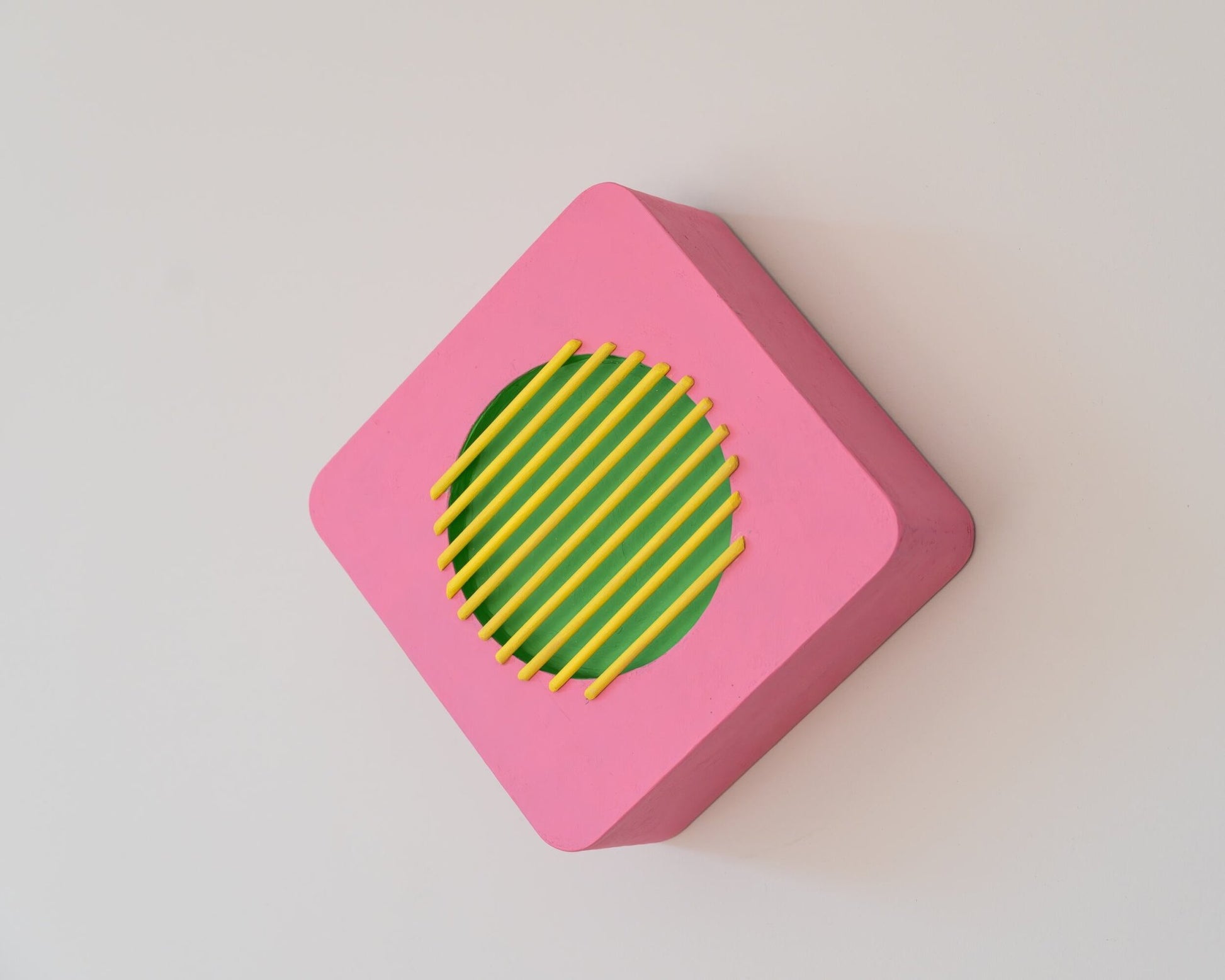 Pink and green geometric wall sculpture displayed on a diagonal, with yellow bars across circular center, highlighting rotatable design.