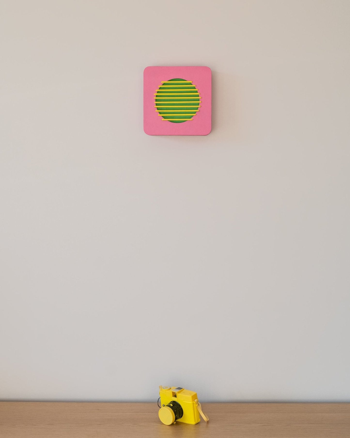 Pink and green geometric wall sculpture mounted above a yellow toy camera on a wooden shelf, highlighting scale and playful interior style.