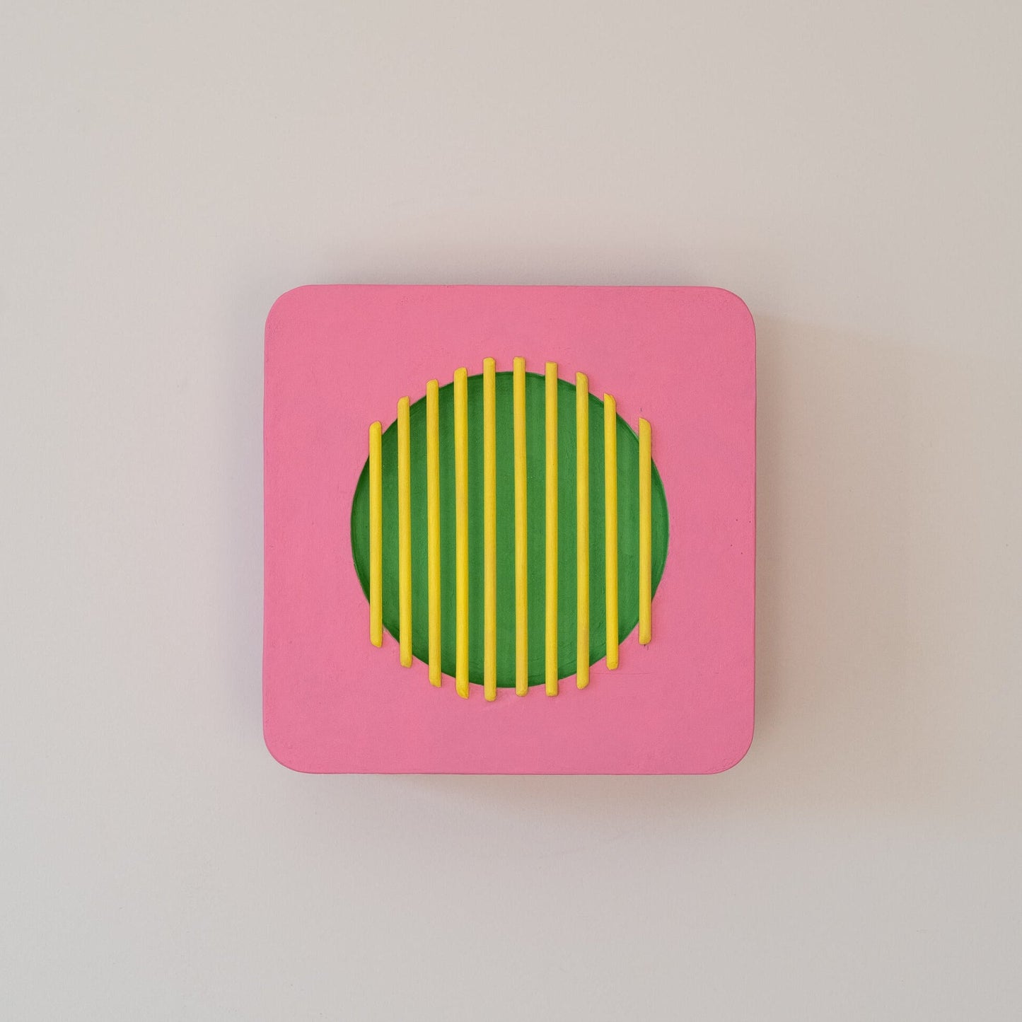 Abstract wall sculpture with pink square frame, green circular center, and yellow vertical bars,
