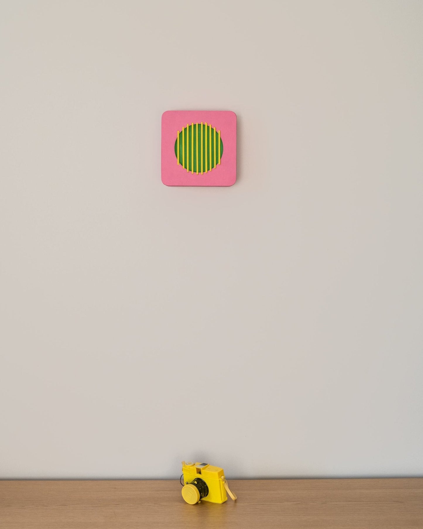 Colorful geometric wall art in pink, green, and yellow displayed above a yellow toy camera on wooden surface, illustrating scale and playful decor.