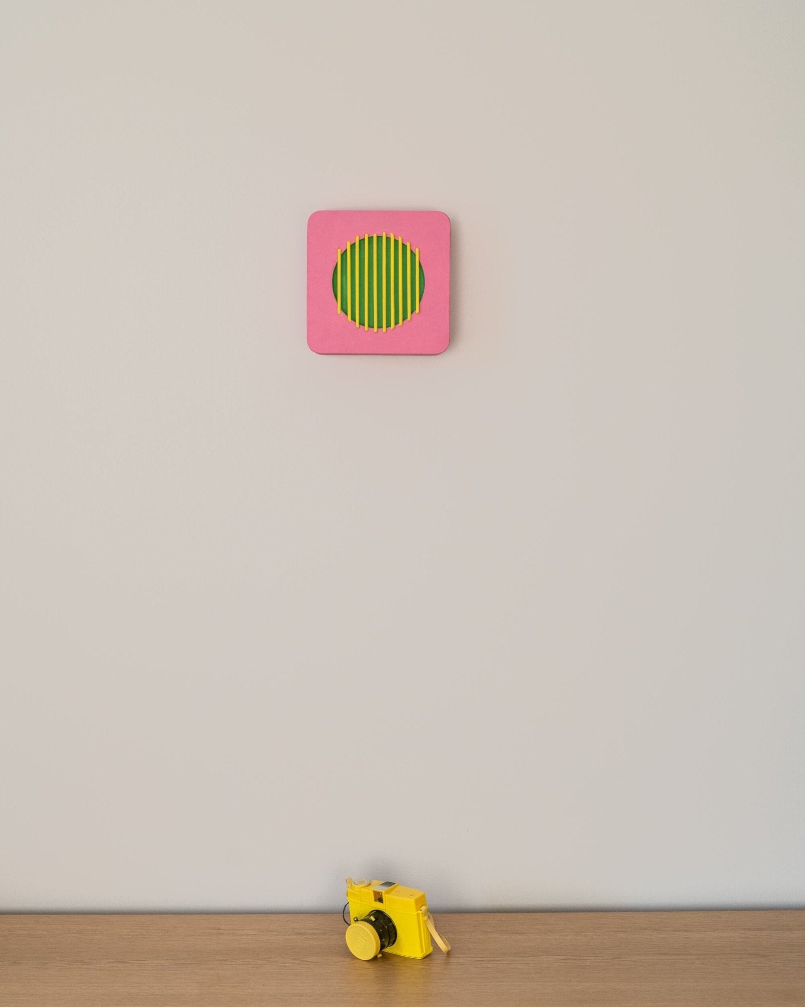 Colorful geometric wall art in pink, green, and yellow displayed above a yellow toy camera on wooden surface, illustrating scale and playful decor.