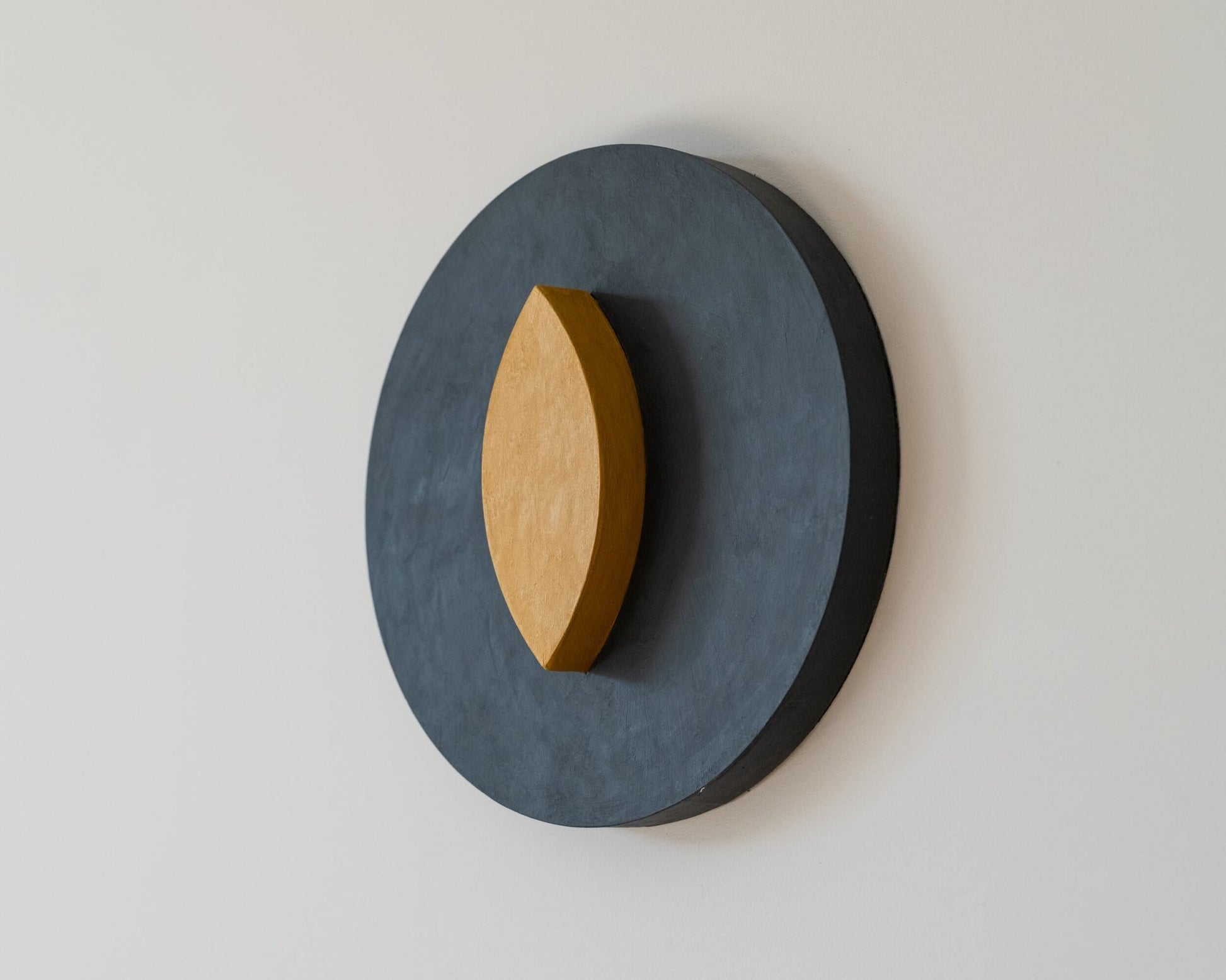3D wall-mounted decorative piece with a dark gray circular base and a  yellow eye shaped oval on a light gray background, 