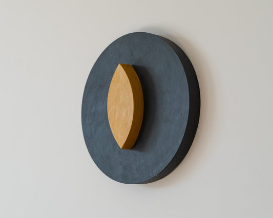 3D wall-mounted decorative piece with a dark gray circular base and a  yellow eye shaped oval on a light gray background, 