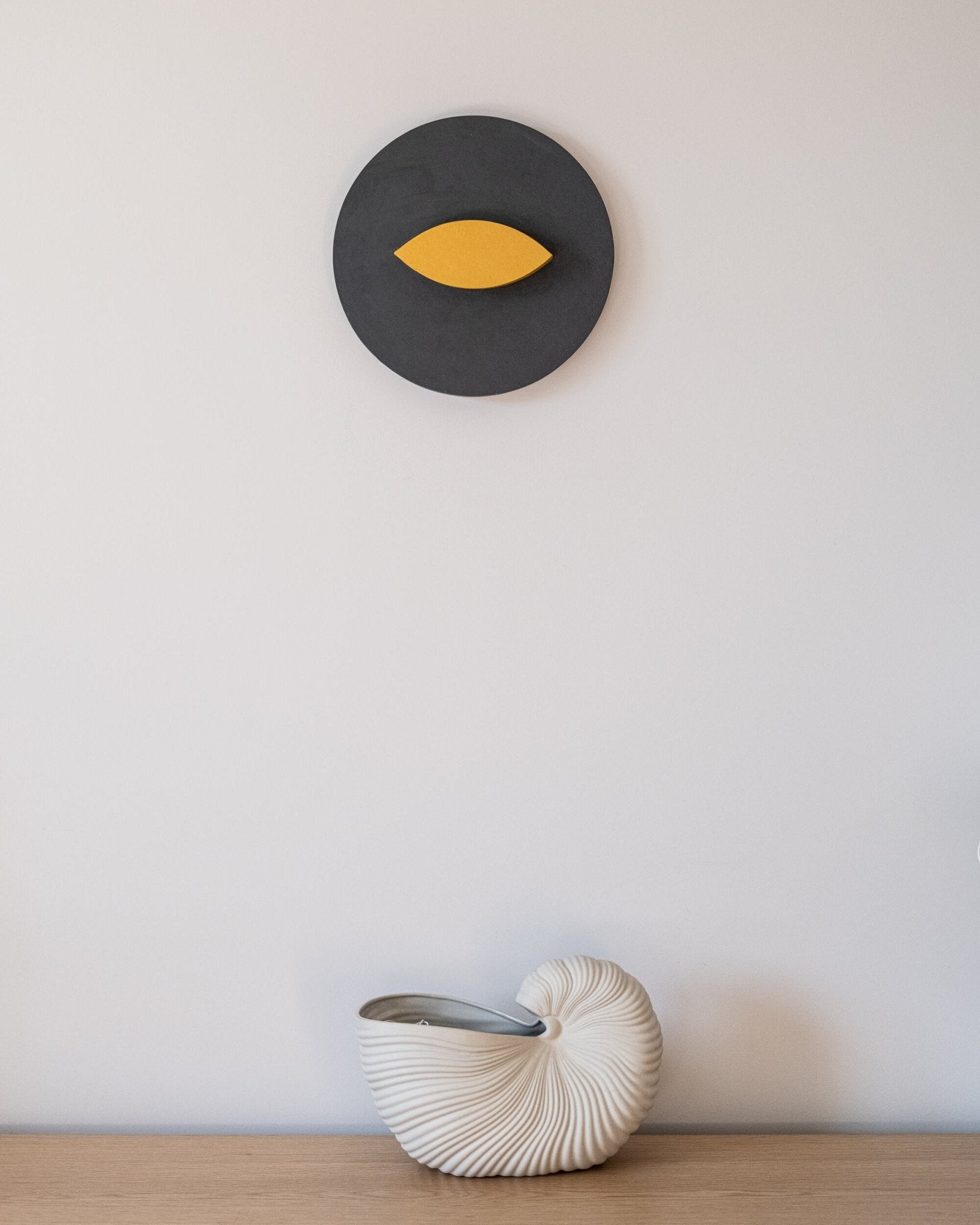 Decorative wall piece with a yellow leaf design on a gray circle, placed above a vase in a shell shape on a wooden surface.