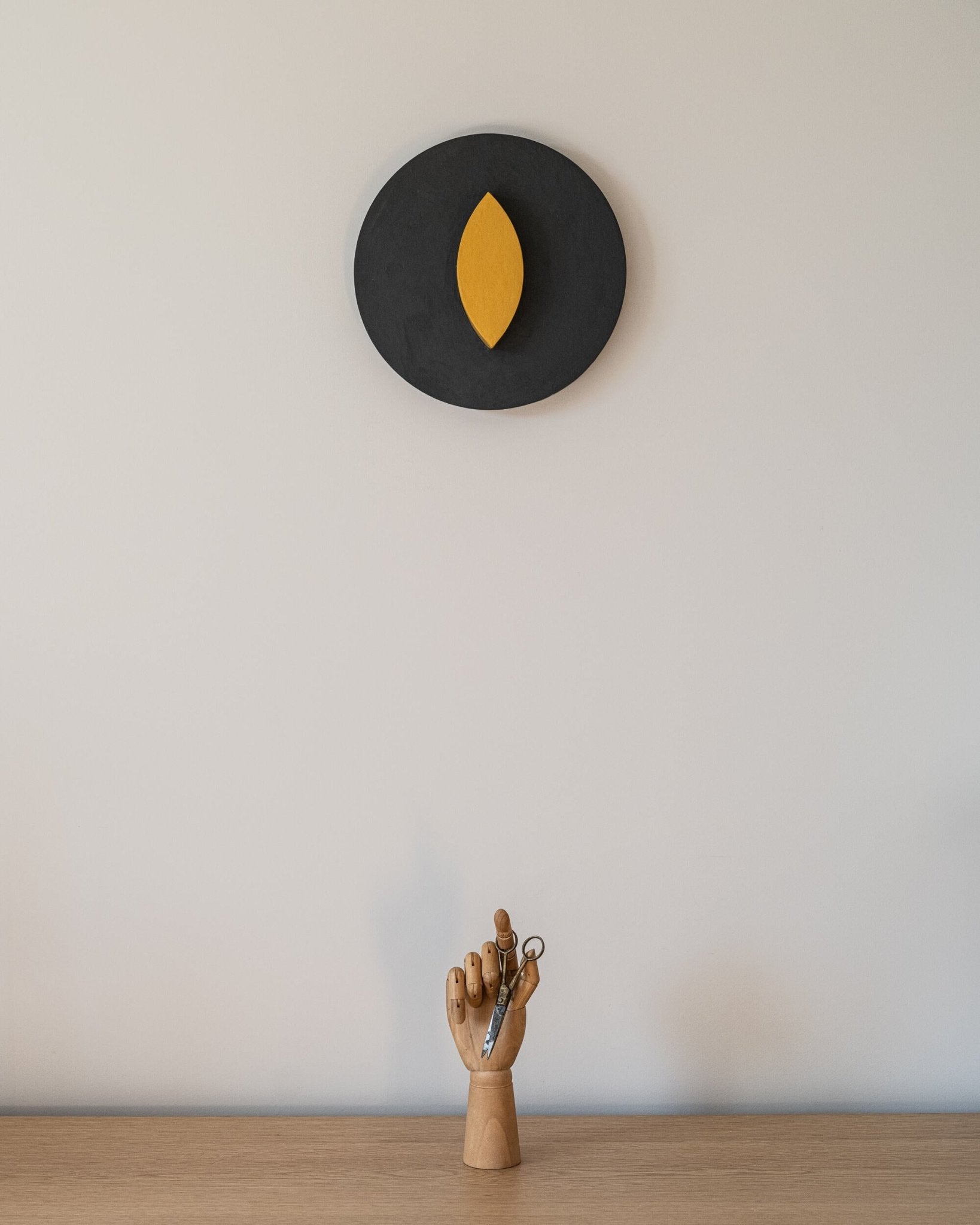 Decorative wall piece with a gray circle and yellow leaf on a plain wall, with a small wooden hand sculpture below.