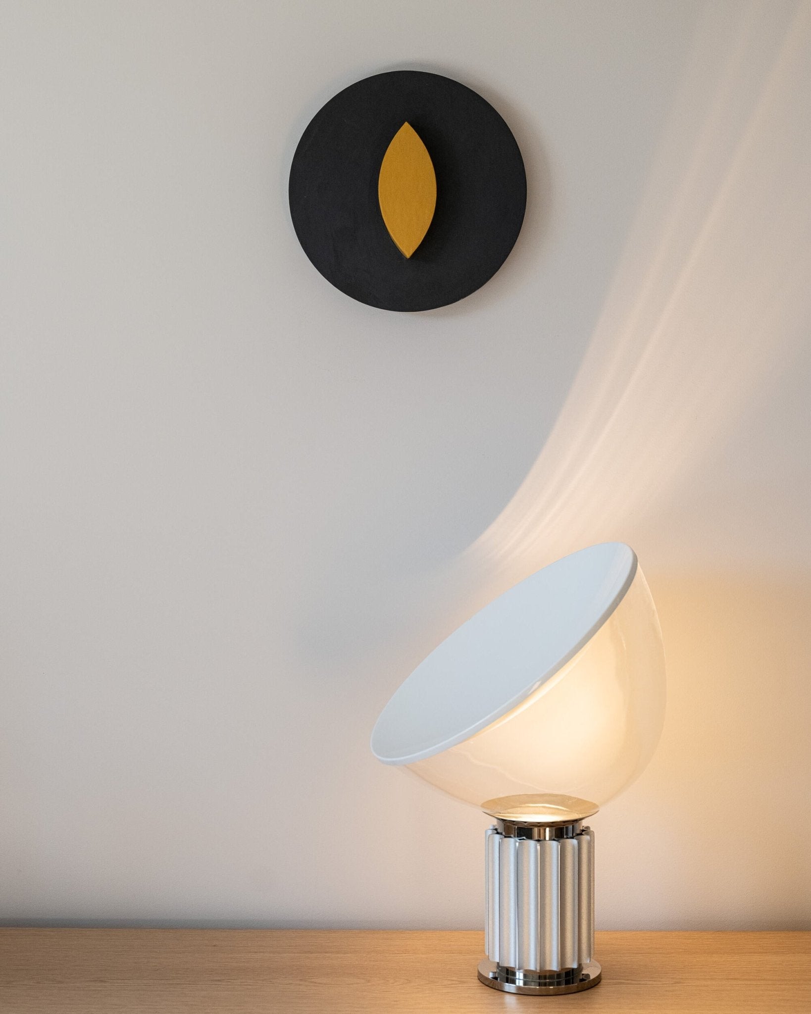 Round wall art object by Olshbau Juicy Center with a modern table lamp with a white shade and metallic base on a wooden surface.