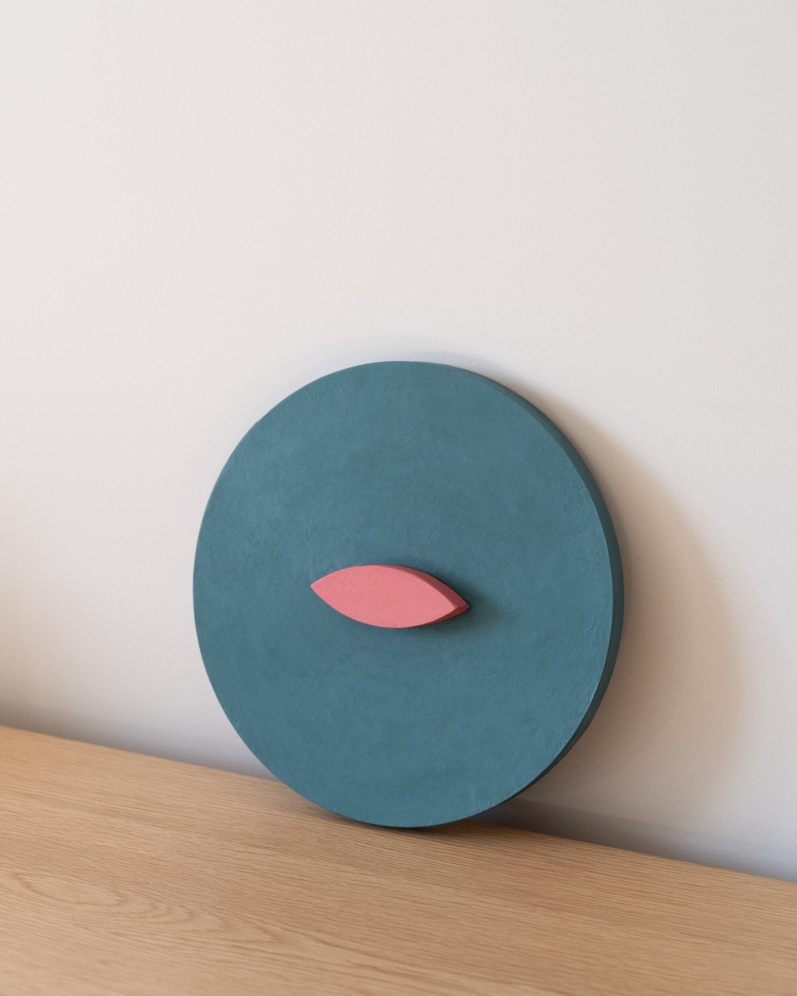 Teal circular 3d art object with a pink leaf-like shape on a light wooden surface and white background