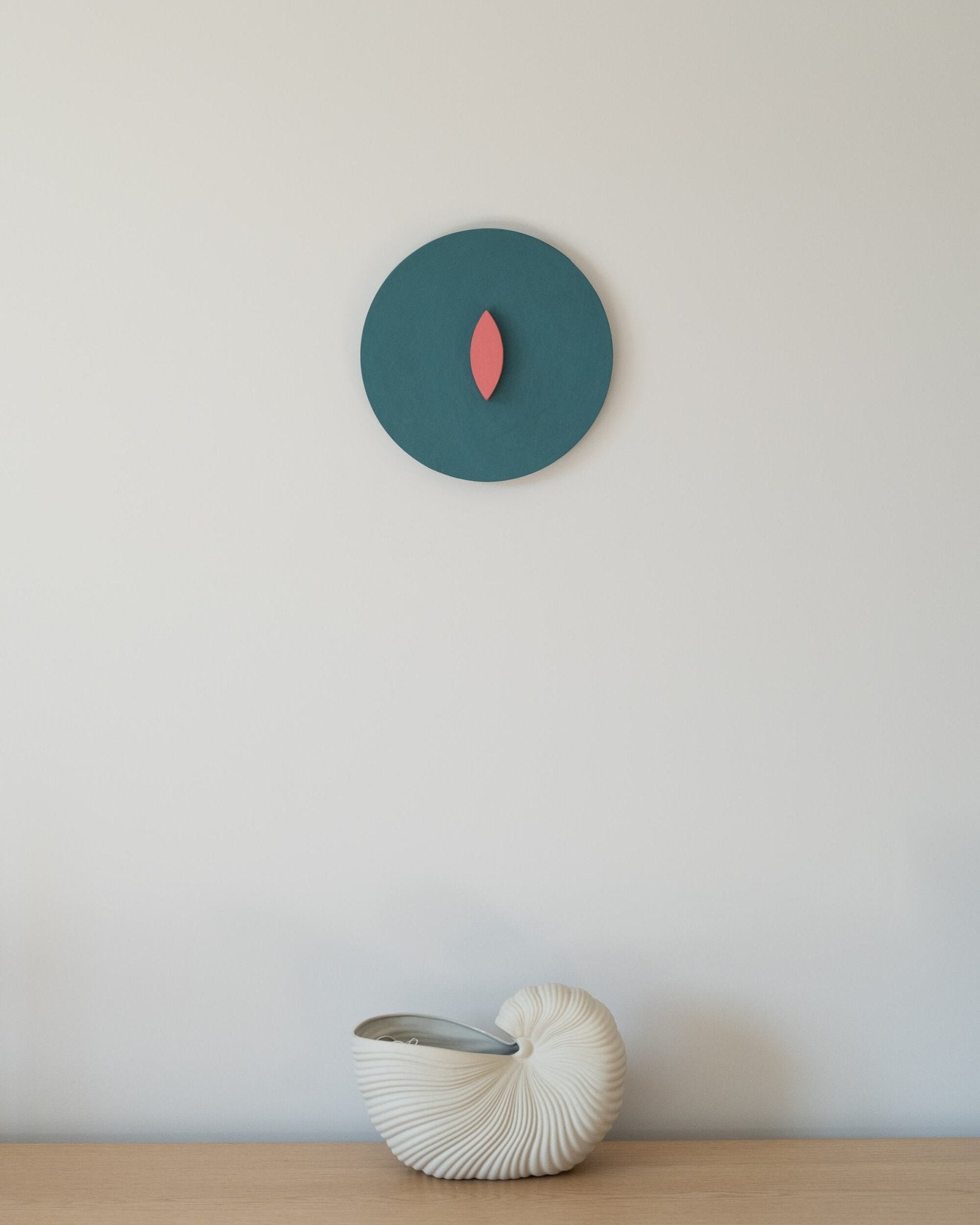 Minimalist round wall sculpture with coral leaf-shaped center on deep teal background, mounted above a white ceramic nautilus vase on a light wooden shelf
