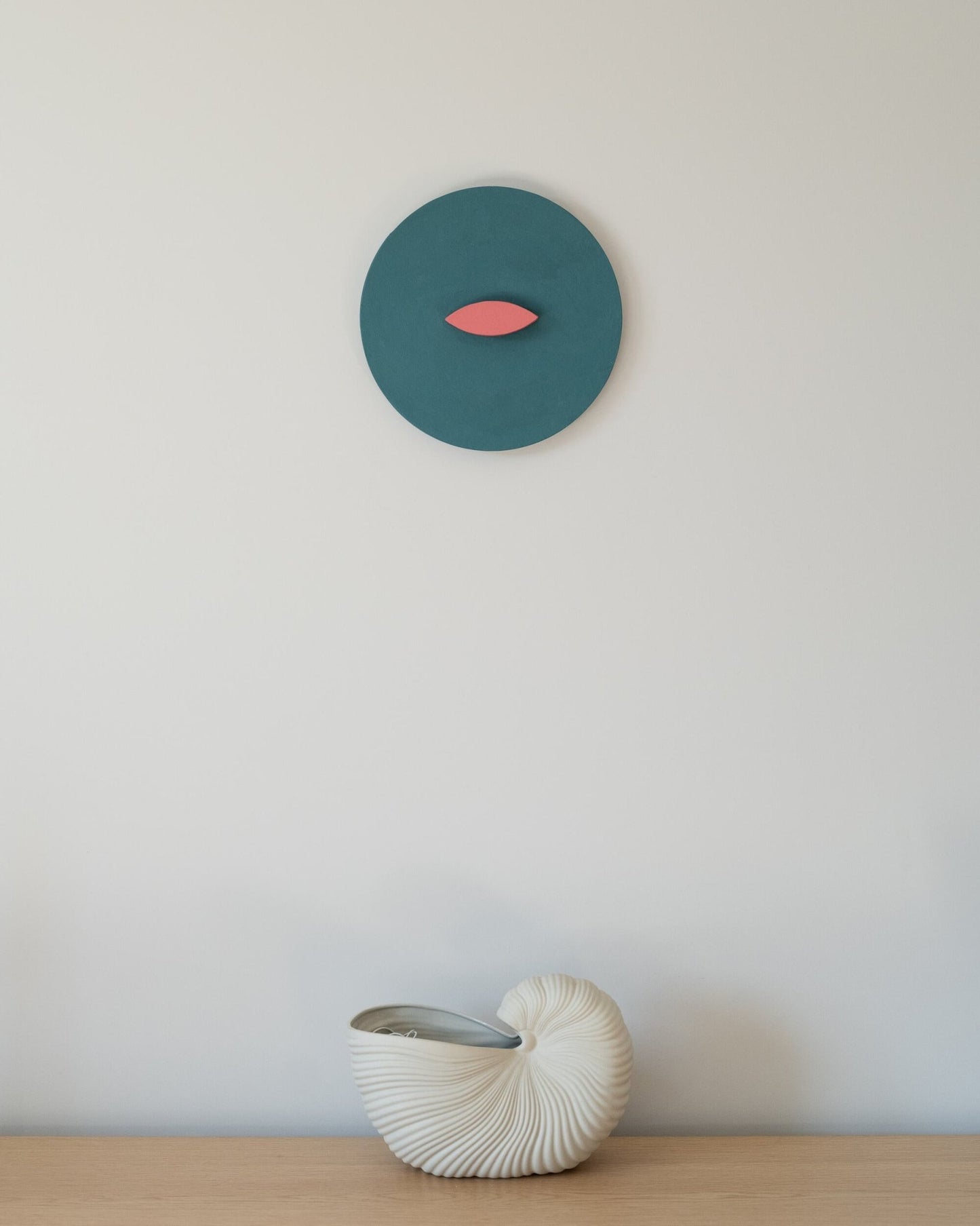 Round blue and coral geometric wall sculpture mounted above a light wooden surface with a white ceramic nautilus vase; minimalist interior styling