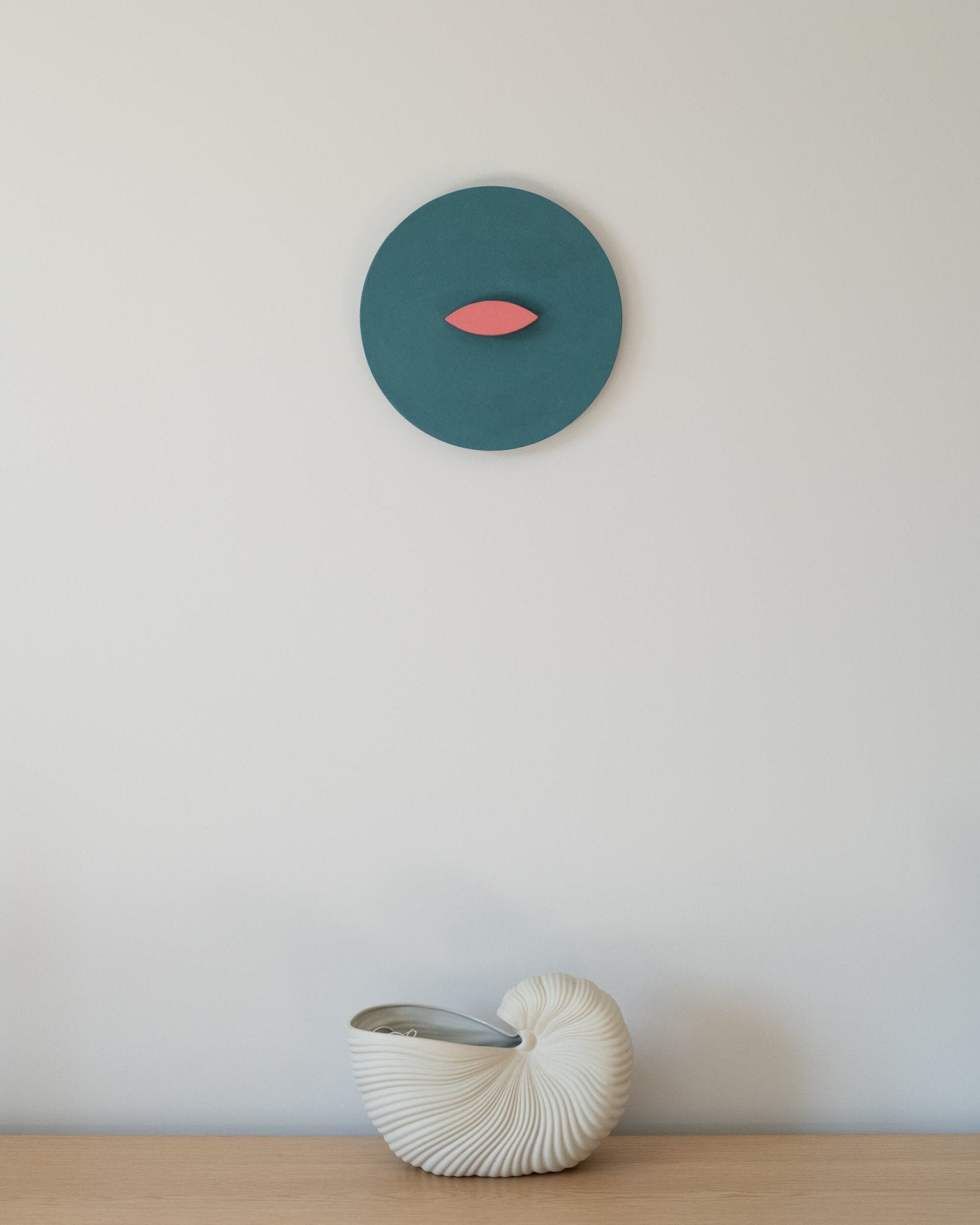 Round blue and coral geometric wall sculpture mounted above a light wooden surface with a white ceramic nautilus vase; minimalist interior styling