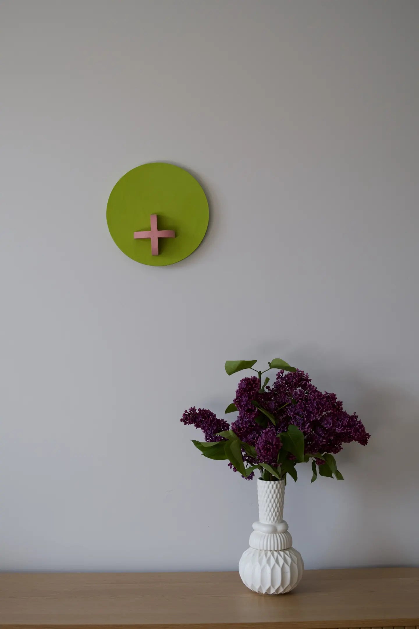 A round green wall sculpture with a pink cross hanging on a white wall above a vase of lilac